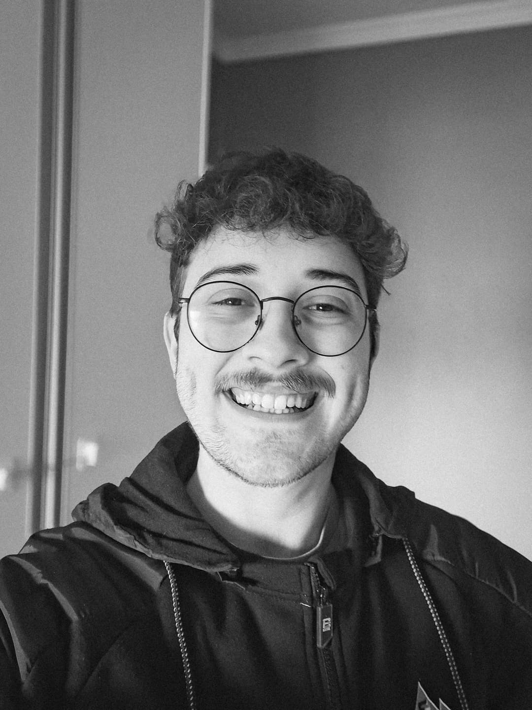 Guilherme Dunguel smiling, wearing rounded glasses and a black jacket, with a blurred background, the photo is black-and-white.